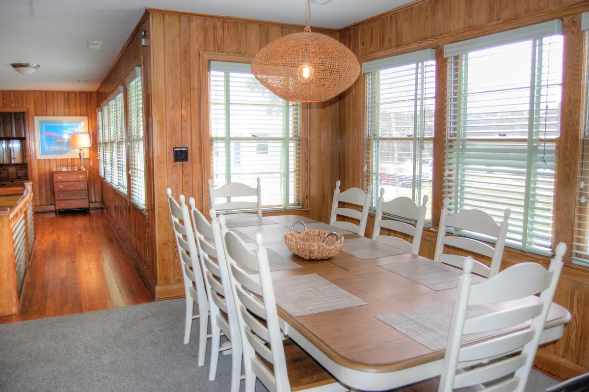 A warm, open view of the adjacent dining area highlighting a long wooden table, woven pendant lighting, and large windows with natural light streaming in. The space is set for family dinners, celebrations, or communal activities such as board games.