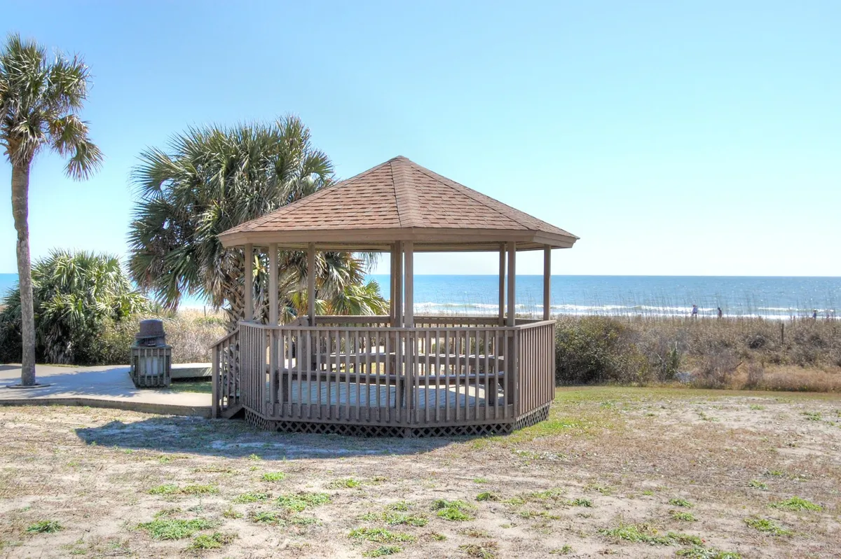 A serene exterior scene featuring a wooden gazebo overlooking the ocean, surrounded by palm trees. The setting invites relaxation and gatherings, with sweeping views of the water and plenty of natural light.