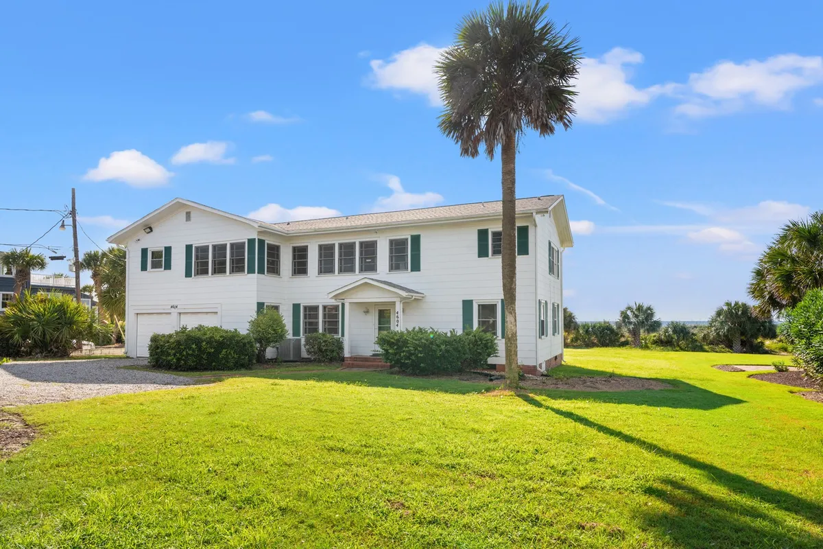 A sun-drenched exterior shot showcasing a charming white house with green shutters, framed by a swaying palm tree in the foreground and an expansive, manicured lawn that sweeps toward the ocean, set against a bright Myrtle Beach sky. The inviting scene hints at ample space for outdoor fun, relaxation, and gatherings.