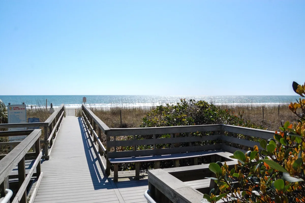 A wooden boardwalk stretches out toward the ocean, crossing dunes and coastal vegetation, with the sea visible in the distance. The image conveys direct and easy access to the beach from the property.
