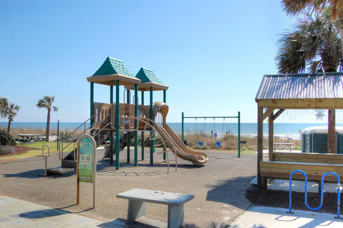 Children are playing on playground equipment with swings, set beside the beach lined with palm trees. The ocean glimmers in the background, and the setting conveys a lively, family-friendly atmosphere.
