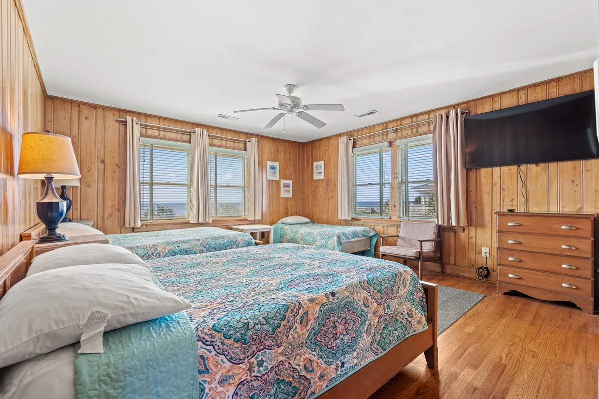 A bright bedroom featuring two beds with colorful bedspreads, wooden furniture, and large windows that bathe the room in natural light. The space appears cheerful and inviting, ideal for family members of all ages.