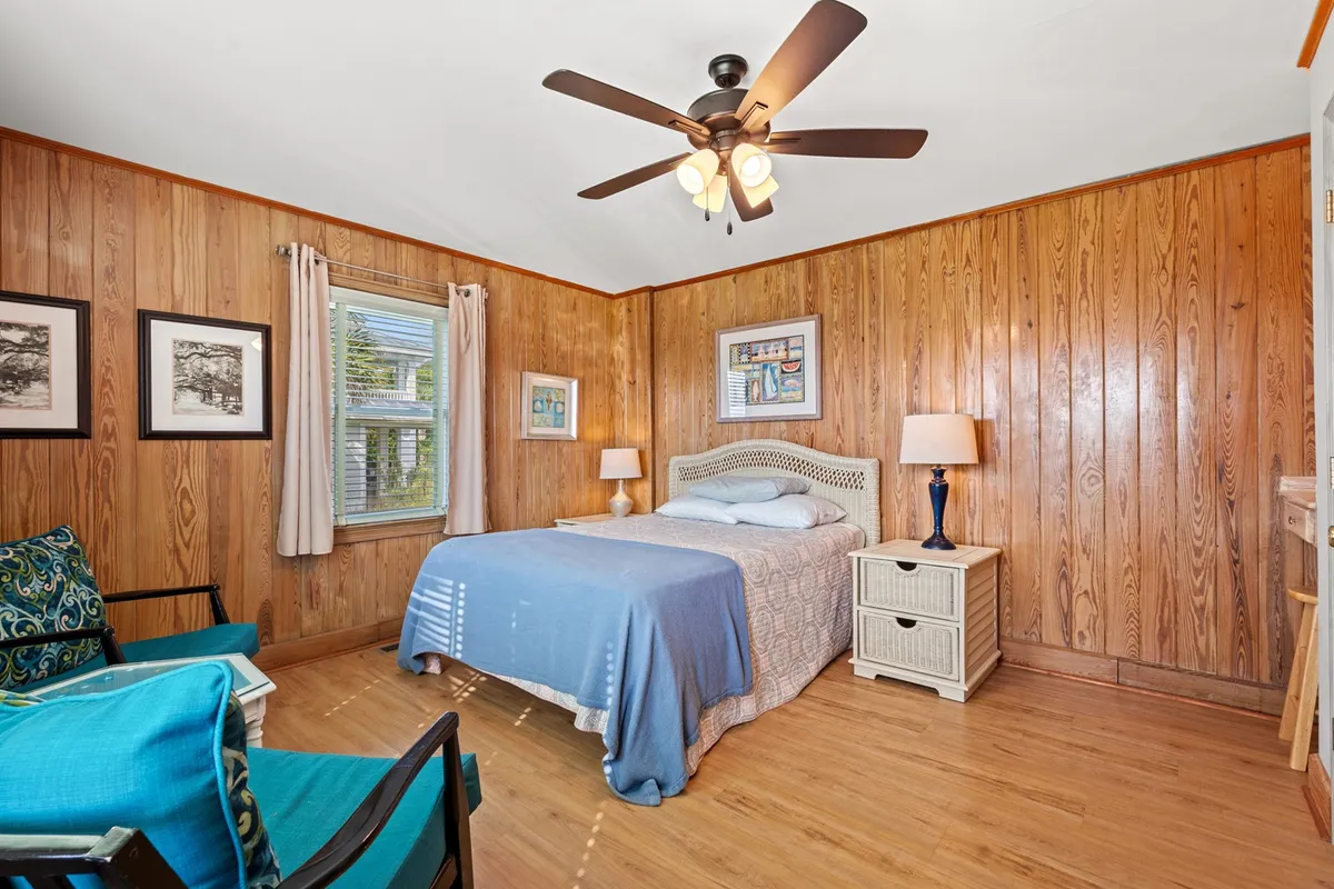 A serene and comfortable bedroom featuring a bed with patterned bedding, a nightstand with a lamp, framed artwork, wood paneling, and a cozy seating area. Sunlight streams in, creating a peaceful retreat for restful nights.