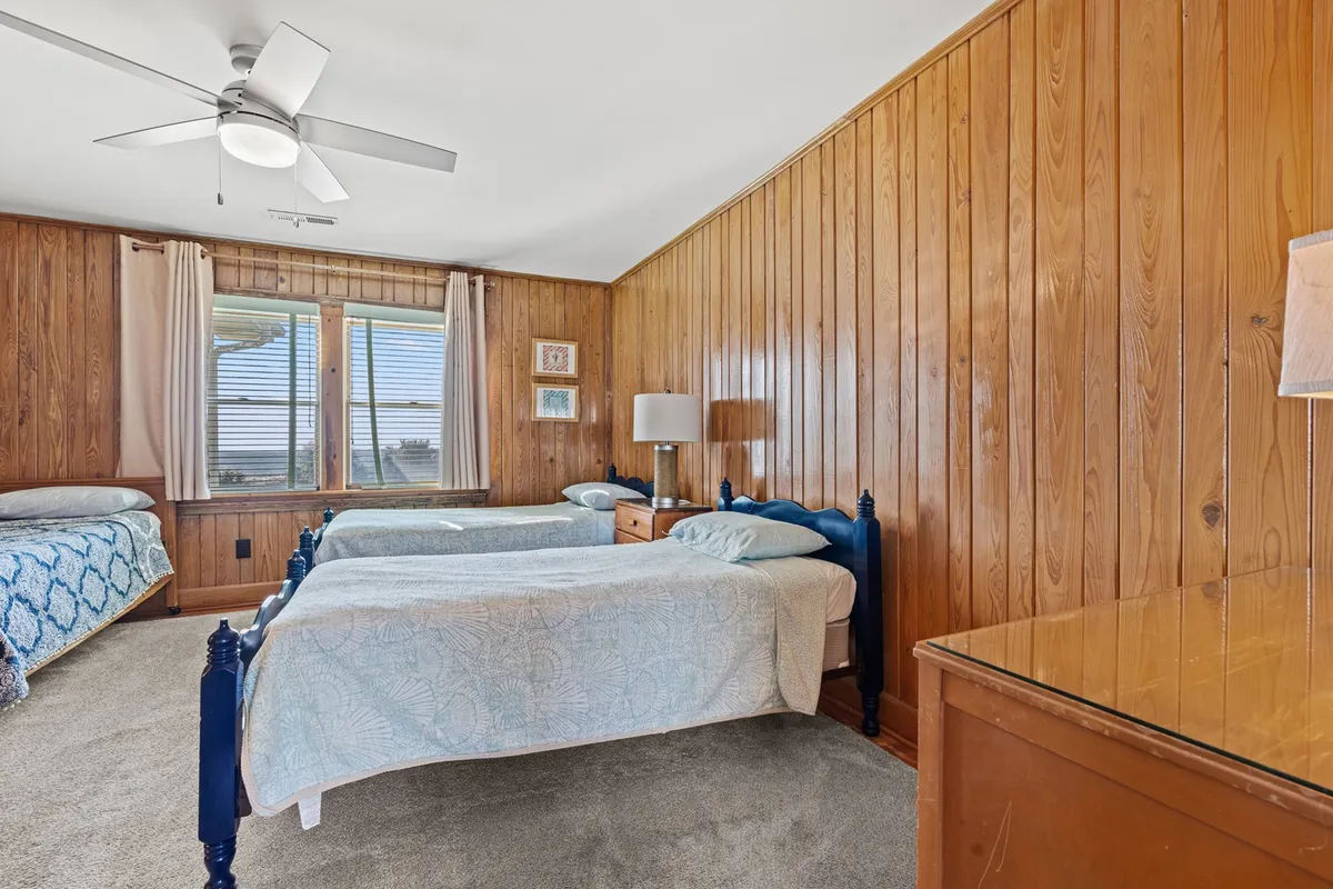A warm, inviting bedroom with twin beds, wooden paneling, a window with curtains, and a lamp on a dresser. The room exudes a relaxed, restful mood, ideal for children or siblings sharing a space during a family vacation.