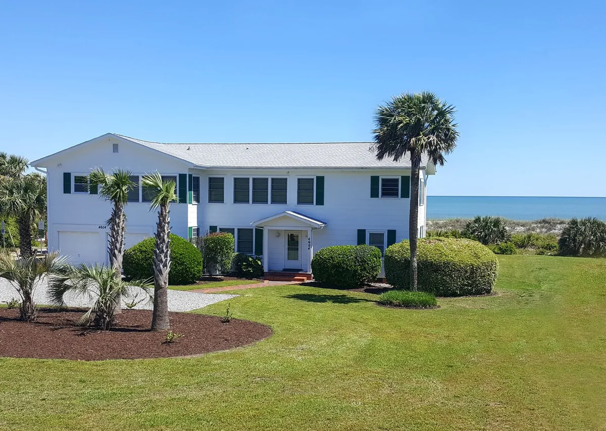 This exterior shot features the house with a wide lawn, palm trees, and a visible sea view in the background. The image showcases the property's spacious private yard and its proximity to the ocean, with the porch offering a welcoming transition between indoor and outdoor living.
