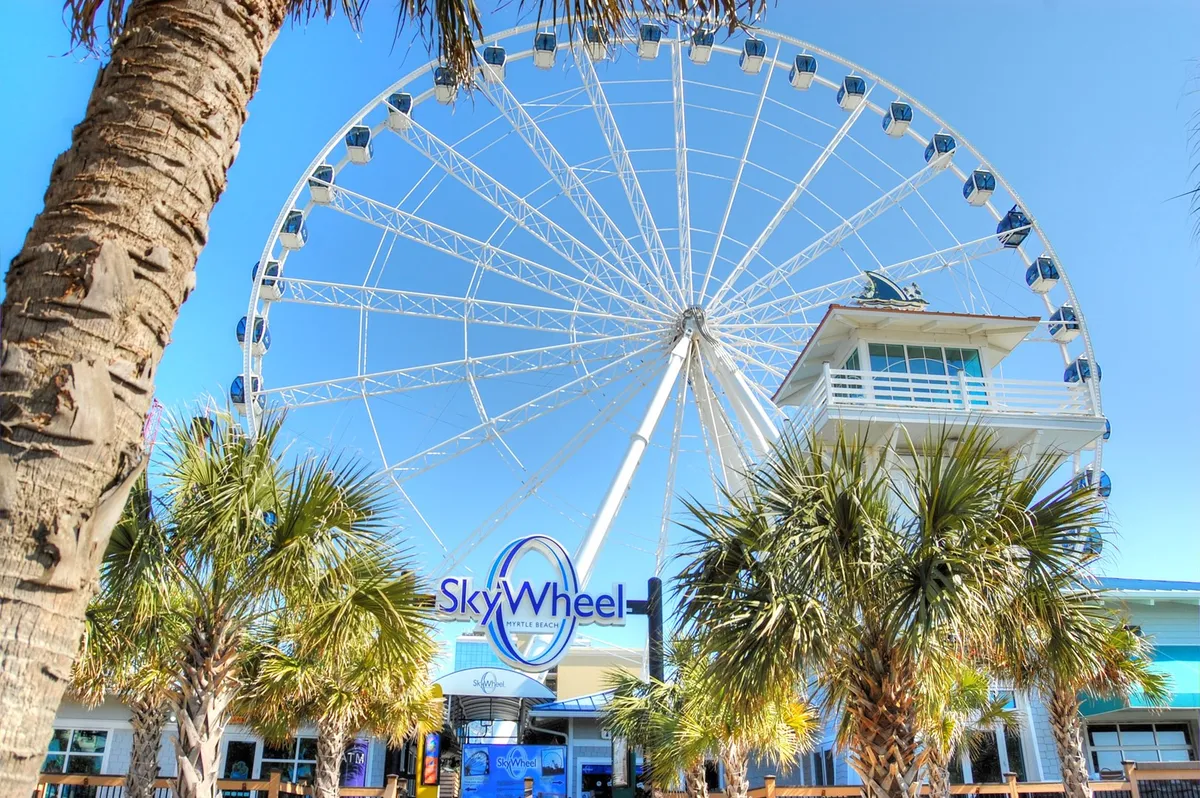 This exterior image features the iconic SkyWheel surrounded by tropical palm trees and a welcoming entrance. The scene is lively and represents one of the top attractions included in the Free Fun Program. The bright, open-air atmosphere captures the excitement and sense of adventure associated with a Myrtle Beach getaway.