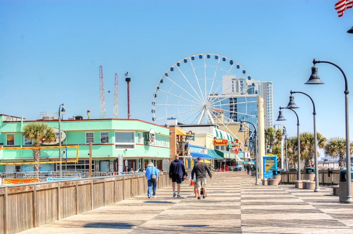 The Myrtle Beach Boardwalk comes alive with a ferris wheel, colorful buildings, palm trees, and bustling activity. The scene radiates fun and excitement, showcasing the variety of attractions just minutes away from the home.